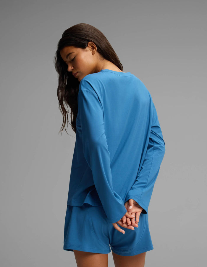 A woman with long dark hair wears the Cozy Earth Women’s First Layer Long Sleeve Sleep Tee in blue, paired with matching shorts. She stands against a plain gray background with her hands clasped behind her back, looking downward.|Color:Anchor