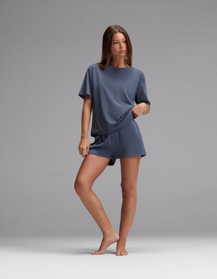 A woman stands barefoot on a gray background, wearing Cozy Earth’s Women’s First Layer Pajama Boxer Short and a matching blue-gray, loose-fitting, short-sleeved top, with one hand in her pocket and the other resting on her thigh.|Color:Dawn