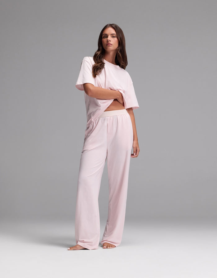 A woman stands barefoot against a gray background, wearing the Cozy Earth Women’s First Layer Pajama Pant in light pink, paired with a loose-fitting, short-sleeved top. Her hair is styled in loose waves and her expression is neutral.|Color:Ballet