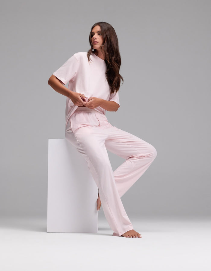 A woman with long brown hair wears the Cozy Earth Women’s First Layer Pajama Pant in light pink and sits barefoot on a white block against a plain gray background.|Color:Ballet