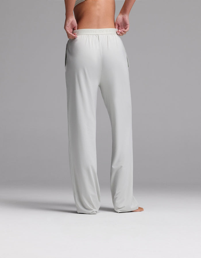 A person stands barefoot on a gray surface, shown from the waist down and back view, wearing Cozy Earth’s Women’s First Layer Pajama Pant in light gray—a loose-fitting style with an elastic waistband and their hands in the pockets.|Color:Laurel