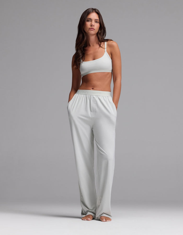 A woman stands barefoot against a plain gray background, wearing a light gray sports bra and Cozy Earth Women’s First Layer Pajama Pants—loose, high-waisted lounge pants with her hands in the pockets.|Color:Laurel