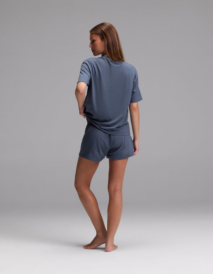 A woman, barefoot and facing away, models the Cozy Earth Women’s First Layer Pajama Short Sleeve Tee & Boxer Short Set in gray against a plain light gray background.|Color:Dawn