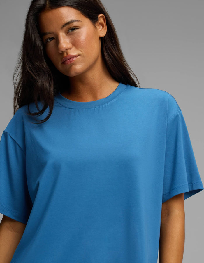 A woman with long dark hair wears the Cozy Earth Women’s First Layer Pajama Short Sleeve Tee in blue, standing against a plain gray background and looking slightly towards the camera with a neutral expression.|Color:Anchor