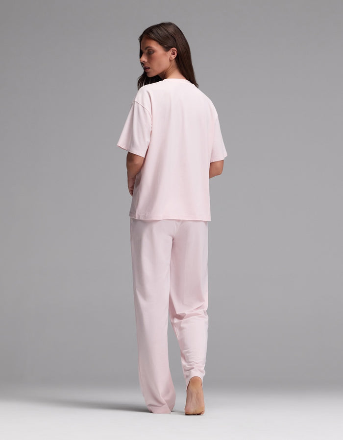 A person stands barefoot on a gray background, facing away from the camera and looking over their shoulder, wearing the Cozy Earth Women’s First Layer Pajama Short Sleeve Tee & Pajama Pant Set in light pink.|Color:Ballet
