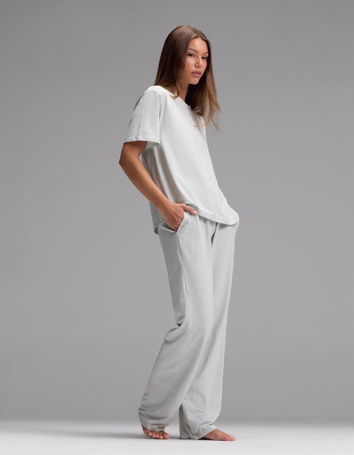 A woman stands barefoot against a gray background, wearing the Cozy Earth Women’s First Layer Pajama Short Sleeve Tee & Pajama Pant Set in light gray, her hands tucked into her pockets as she gazes slightly to the side.|Color:Laurel