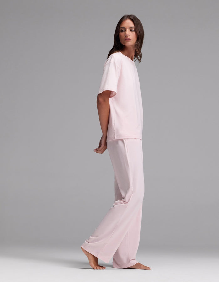 A woman with long brown hair wears the Cozy Earth Women’s First Layer Pajama Short Sleeve Tee & Pajama Pant Set in light pink. She stands barefoot on a plain gray background, looking over her shoulder with a neutral expression.|Color:Ballet