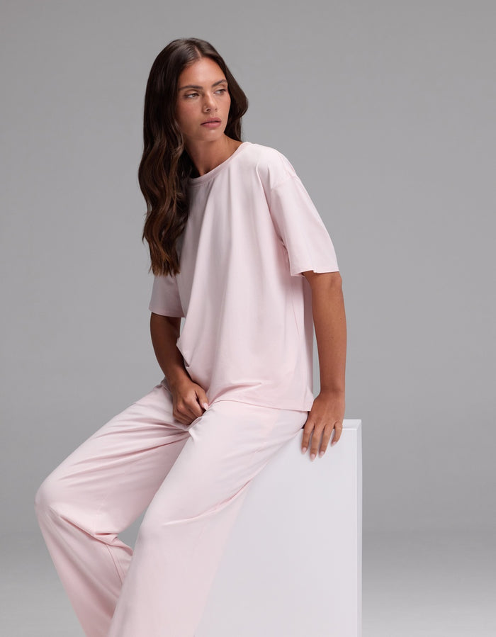 A woman with long brown hair is wearing the Cozy Earth Women’s First Layer Pajama Short Sleeve Tee & Pajama Pant Set in light pink as she sits on a white block, looking to the side against a plain gray background.|Color:Ballet