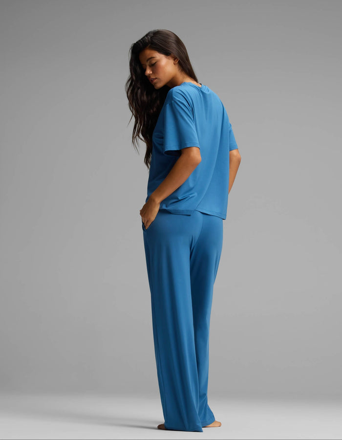 A woman stands barefoot, facing sideways and looking over her shoulder. She is wearing the Cozy Earth Women’s First Layer Pajama Short Sleeve Tee & Pant Set in blue against a plain grey background.|Color:Anchor