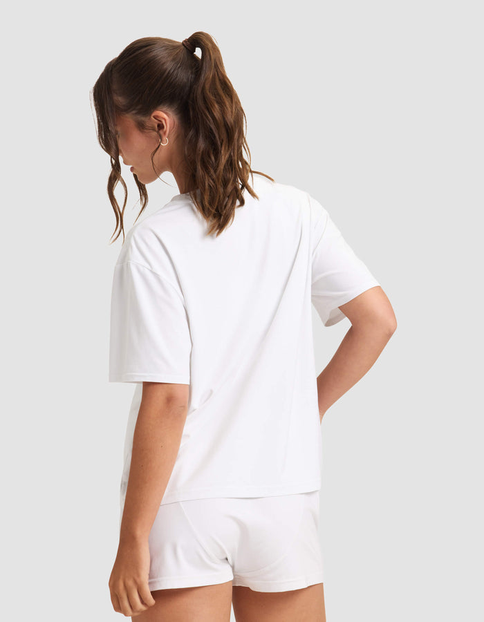 A woman with wavy brown hair in a ponytail stands with her back to the camera, wearing Cozy Earth's Women's First Layer Pajama Short Sleeve Tee and white shorts against a plain light gray background.|Color:White