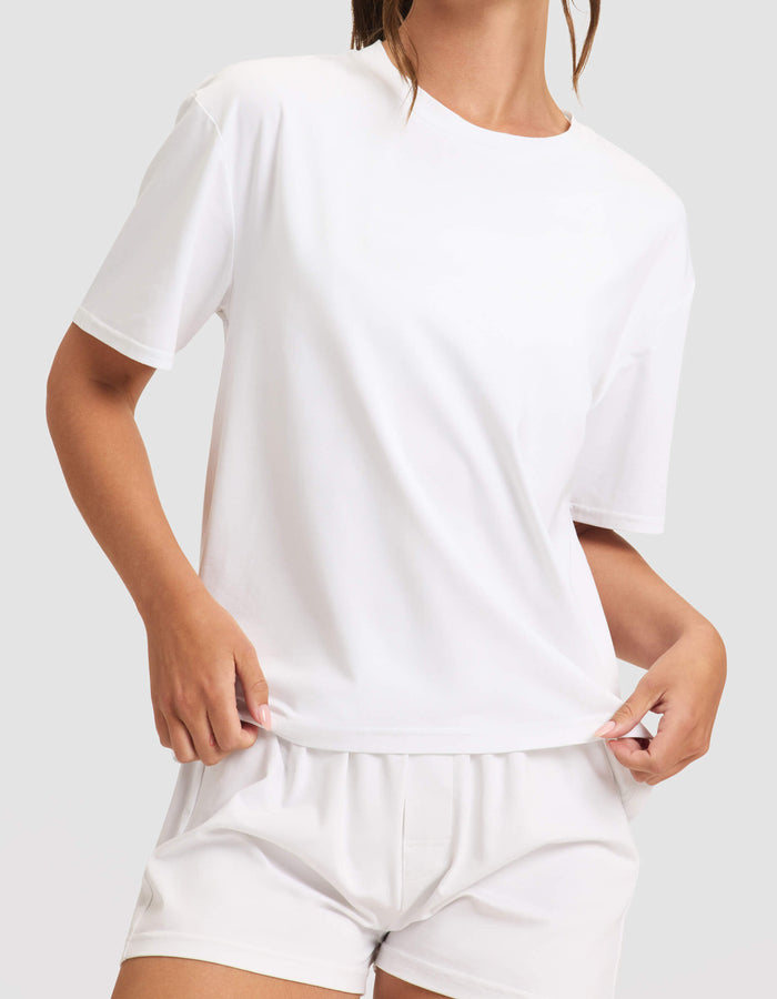 A woman wears the Cozy Earth Women's First Layer Pajama Short Sleeve Tee and matching white shorts, standing against a neutral background while holding the shirt's hem with one hand.|Color:White