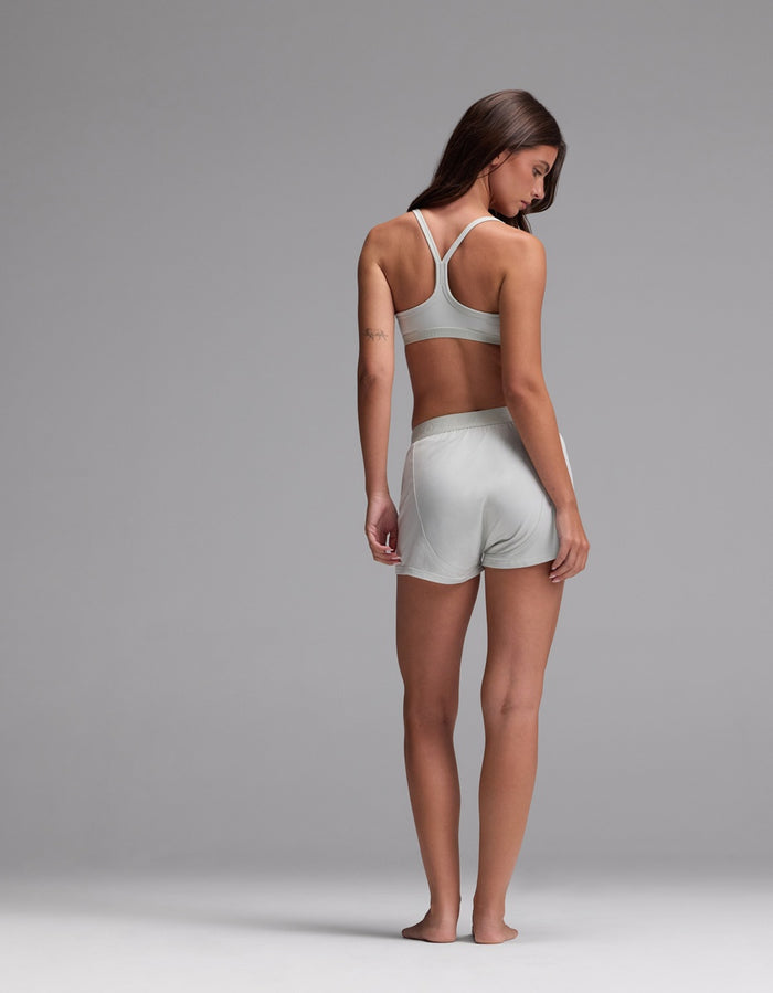 A woman stands barefoot on a gray background, facing away while wearing the Cozy Earth Women’s First Layer Pajama Soft Bra & Boxer Short Set. She looks over her shoulder with her arms relaxed by her sides.|Color:Laurel