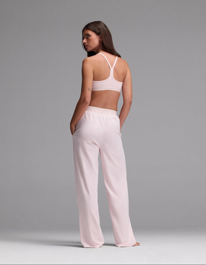 A woman stands with her back to the camera, wearing the Cozy Earth Women’s First Layer Pajama Soft Bra & Pajama Pant Set in light pink. Her hands rest in her pockets as she looks over her shoulder against a plain gray background.|Color:Ballet