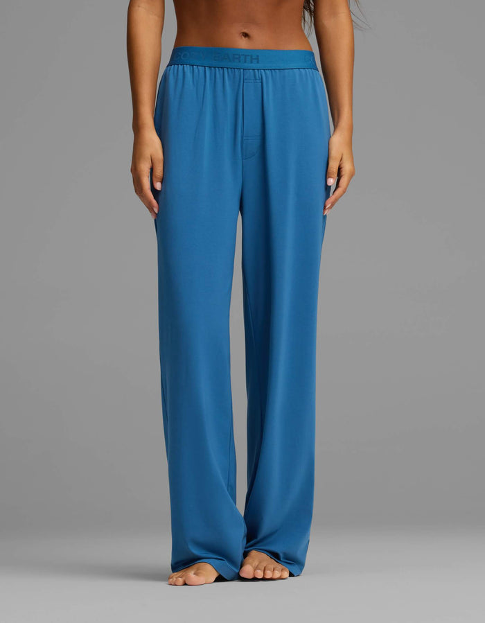 A person is shown barefoot, wearing the Cozy Earth Women’s First Layer Pajama Short Sleeve Tee & Pant Set in blue. The image focuses on the lower torso and legs, highlighting the relaxed, high-waisted pants with an elastic waistband against a plain gray background.|Color:Anchor