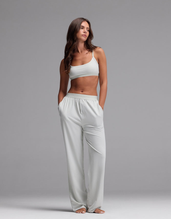 A woman stands barefoot against a gray background, wearing the Cozy Earth Women’s First Layer Soft Bra paired with matching loose pants. Her hands are in her pockets and she gazes slightly to the side with a neutral expression.