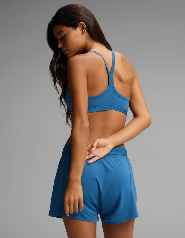 A woman with long dark hair stands in a relaxed pose—one hand behind her back—wearing the Cozy Earth Women’s First Layer Soft Bra and matching shorts against a plain gray background.|Color:Anchor