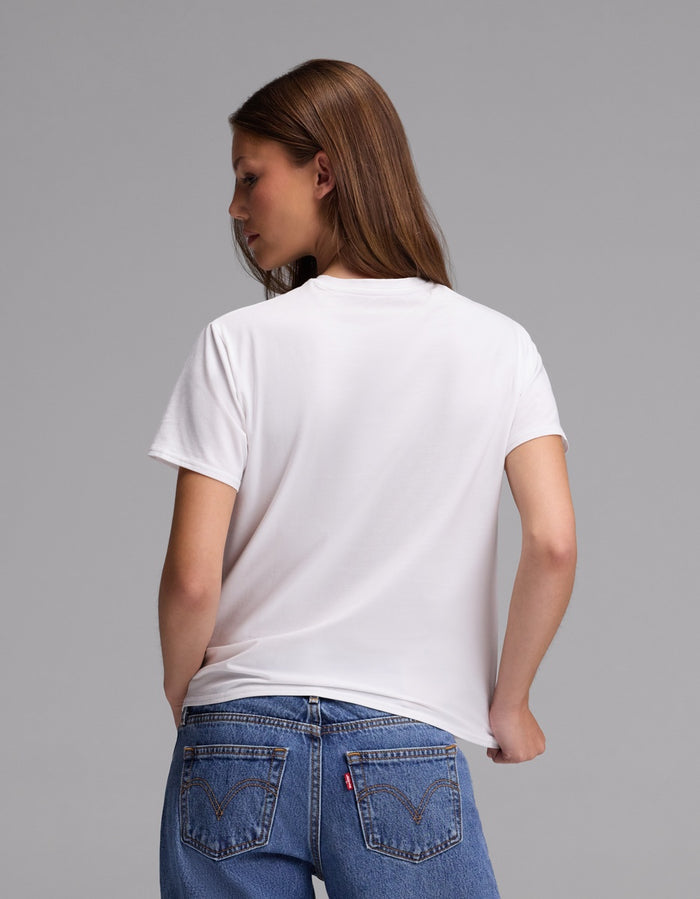A woman with long brown hair is seen from behind, standing against a gray background while wearing the Cozy Earth Women’s Go-To Tee in white paired with blue jeans.|Color:White