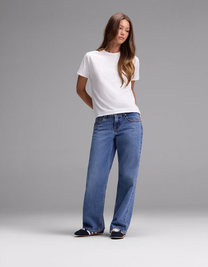 A person stands against a plain gray background, wearing the Cozy Earth Women’s Go-To Tee in white, blue wide-leg jeans, and black slip-on shoes with white accents.|Color:White