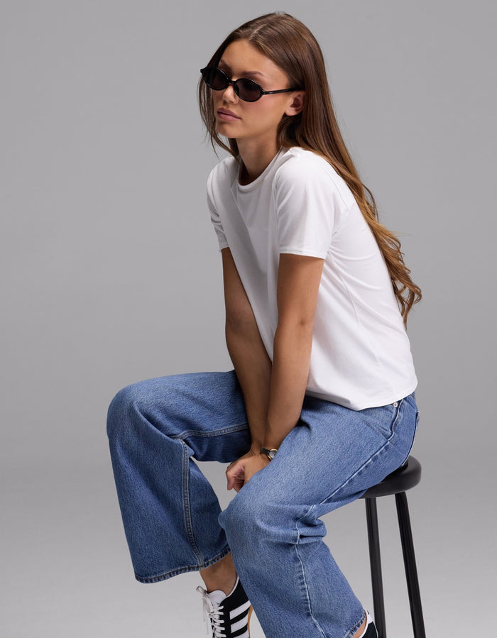A person with long brown hair wears sunglasses, blue jeans, and black sneakers, sitting on a black stool against a plain gray background. She is wearing the Cozy Earth Women’s Go-To Tee.|Color:White