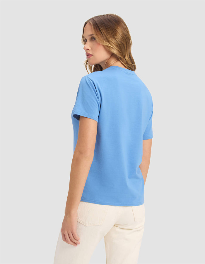 A woman with light brown hair wears the Cozy Earth Women's Go-To Tee in light blue and off-white pants, standing against a plain light gray background, facing away while turning her head slightly to the left.|Color:Sailor