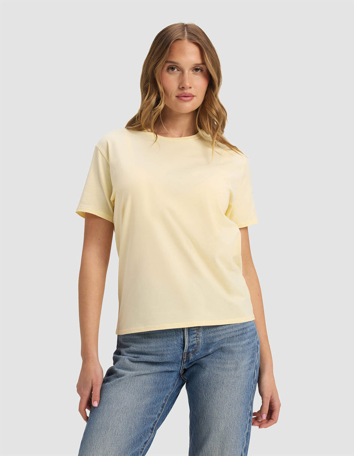 A woman with wavy light brown hair wears the Cozy Earth Women's Go-To Tee in Meringue and blue jeans, standing against a plain light gray background.|Color:Meringue
