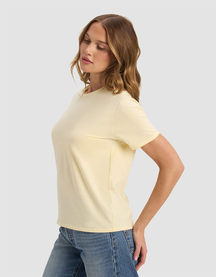 A woman with wavy, light brown hair wears the Cozy Earth Women's Go-To Tee in pale yellow and blue jeans. She stands sideways, eyes closed, arms slightly behind her against a plain light background.|Color:Meringue