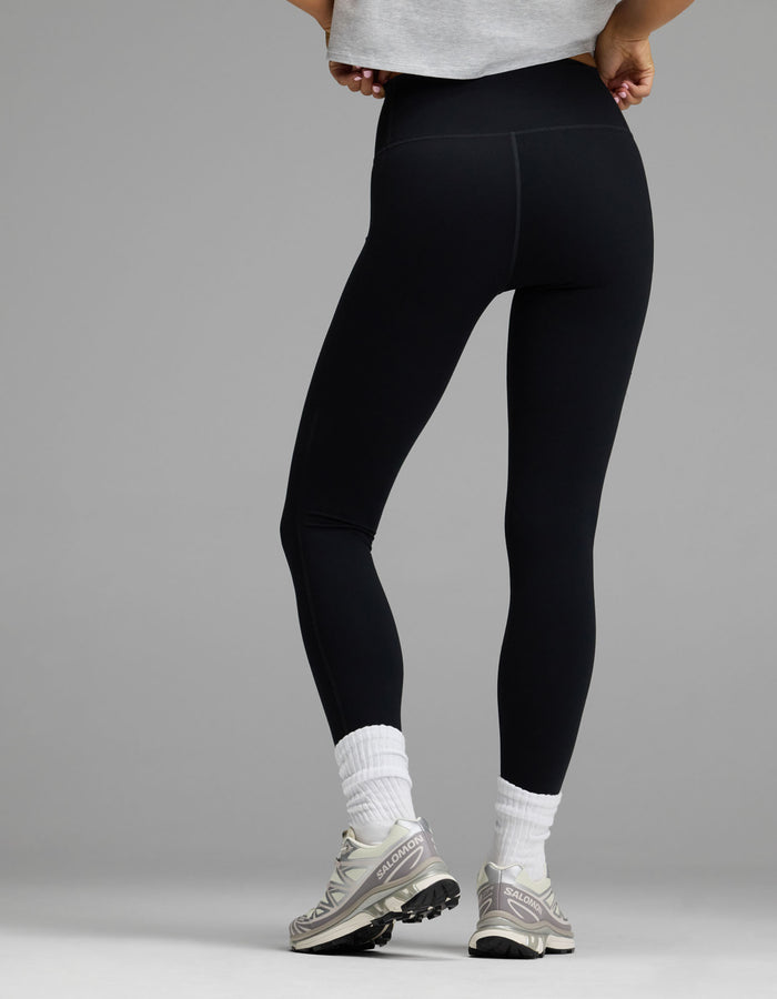 A person stands with their back to the camera wearing Cozy Earth’s Women’s Liv Legging, paired with white socks, gray sneakers, and a light gray cropped shirt, set against a plain gray background.|Color:Jet Black