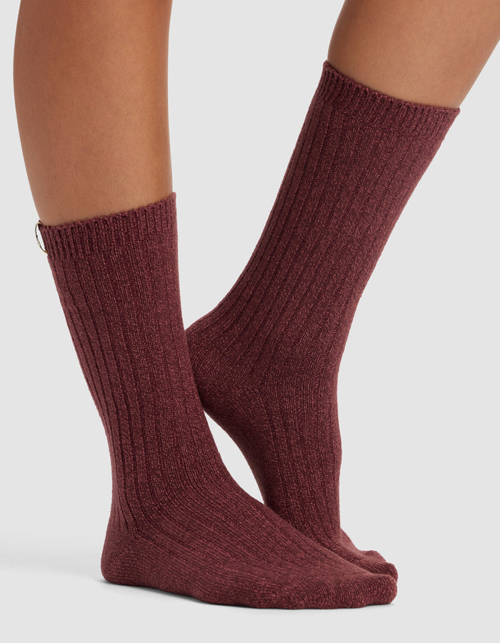 A pair of bare legs wears The Plush Lounge Calf Sock from Cozy Earth in ribbed, dark mauve, mid-calf length, featuring a snug fit and subtle texture, standing on a light surface.|Color:Burgundy/Rouge/Dusty Rose