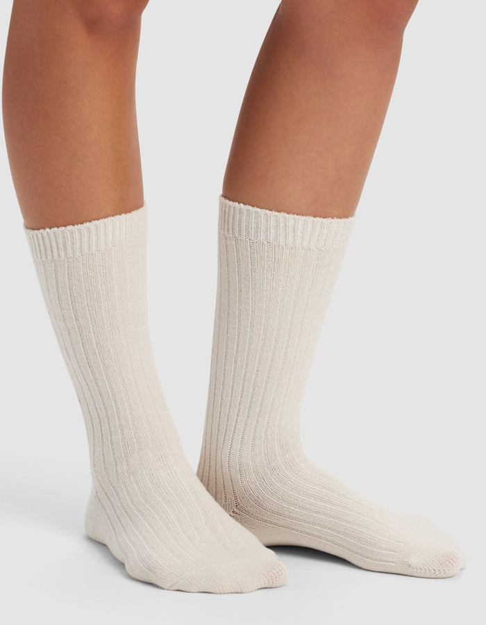 Someone wearing Cozy Earth's The Plush Lounge Calf Sock 3-Pack in a thick, cream-colored ribbed style that reaches mid-calf, standing on a white surface with only their lower legs and feet visible.|Color:Cacao/Taupe/Alabaster