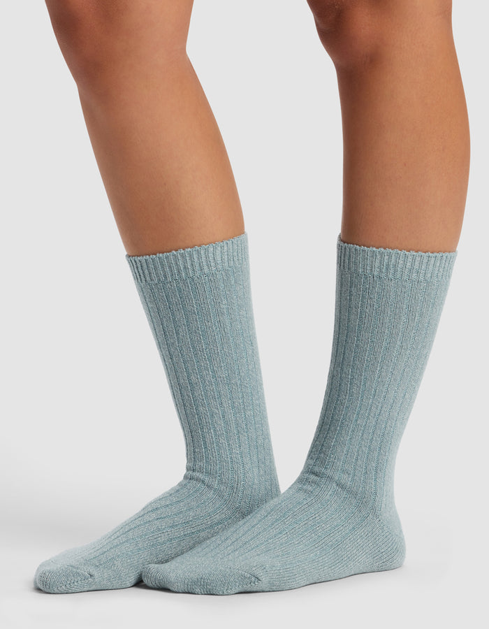 Someone wearing Cozy Earth’s The Plush Lounge Calf Sock 3-Pack in light blue, featuring a ribbed crew design, stands on a white surface, displaying their lower legs and feet.|Color:Eclipse/Deep Blue/Smokey Blue