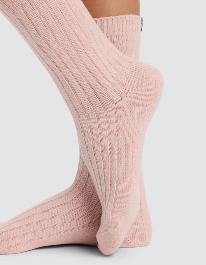 Close-up of two feet wearing light pink Cozy Earth The Plush Lounge Calf Sock 3-Pack socks. One foot is flat on a plain light gray background, while the other is elevated, resting on the toes.|Color:Burgundy/Rouge/Dusty Rose