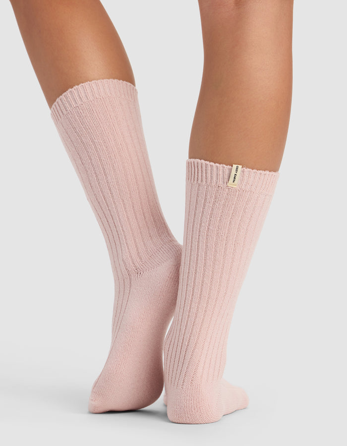 A person models Cozy Earth’s The Plush Lounge Calf Sock 3-Pack in light pink ribbed style, standing on a light surface with ankles crossed. A small fabric tag is visible near the top of one sock.|Color:Burgundy/Rouge/Dusty Rose