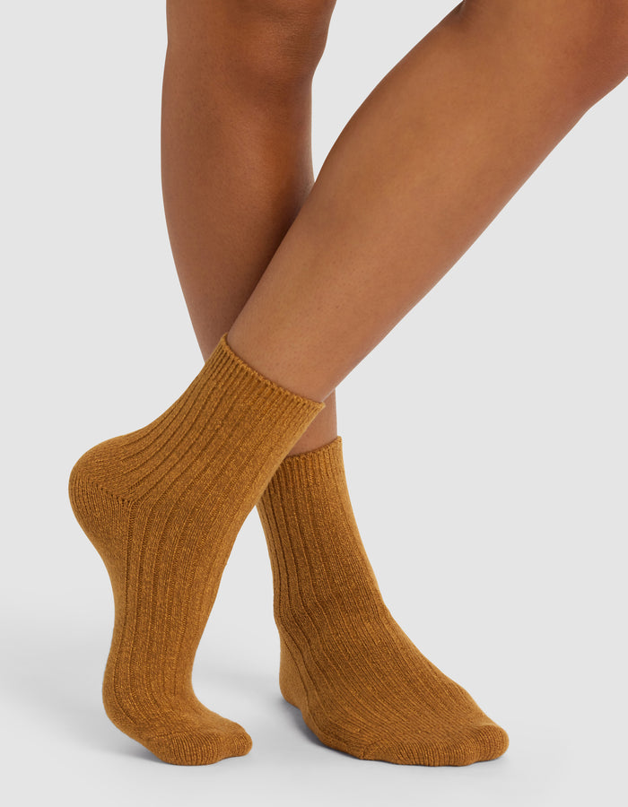 A person stands with legs crossed wearing Cozy Earth's Plush Lounge Quarter Socks 3-Pack in a thick, ribbed, mustard-colored style on a white background.|Color:Nubuck/Amber/Daisy
