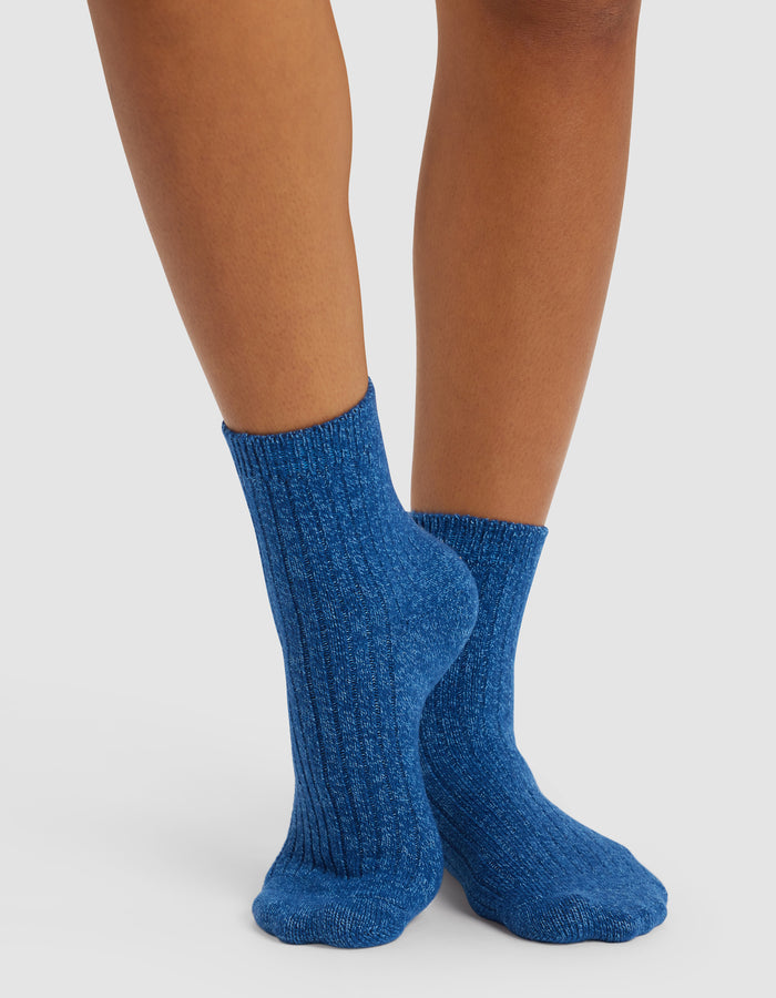 Someone wearing Cozy Earth’s Plush Lounge Quarter Sock 3-Pack in blue stands on a light surface with their feet crossed, showcasing the socks’ textured ribbed design.|Color:Pacific/Harbor/Lake