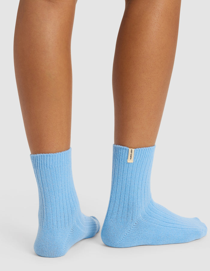 A person wears Plush Lounge Quarter Sock 3-Pack by Cozy Earth in light blue, featuring a small beige label on the back. Only the lower legs and feet are shown against a white background.|Color:Pacific/Harbor/Lake