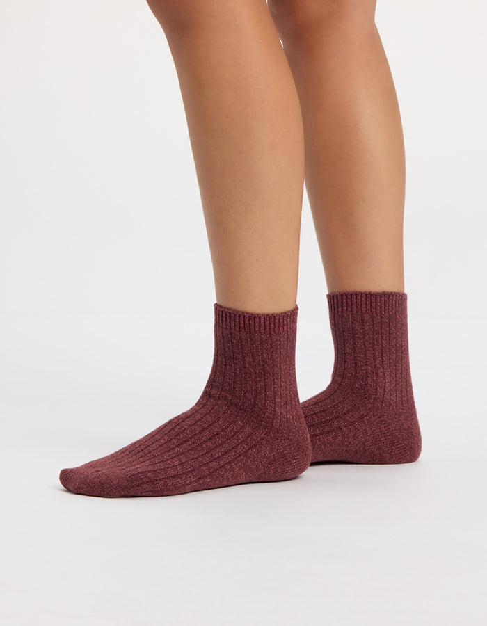 A person wearing maroon socks from the Cozy Earth Plush Lounge Quarter Sock 3-Pack stands on a white surface, with only their lower legs and feet visible.|Color:Burgundy/Tulip/Blossom