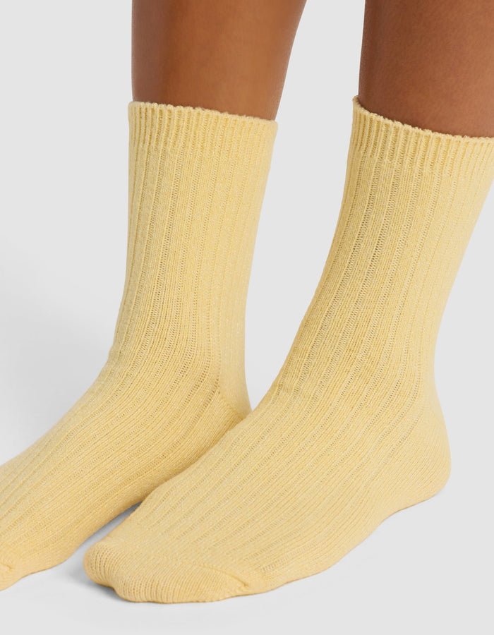 A close-up of lower legs and feet wearing Cozy Earth's The Plush Modern Crew Sock in light yellow, standing on a white surface.|Color:Daisy