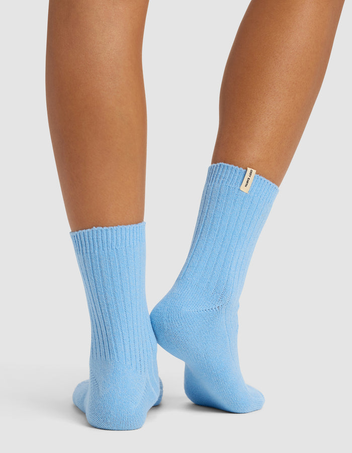 A pair of legs stands on tiptoes, showing the backs of ribbed, light blue Cozy Earth Plush Modern Crew Socks with a small beige tag on one sock against a plain white background.|Color:Harbor