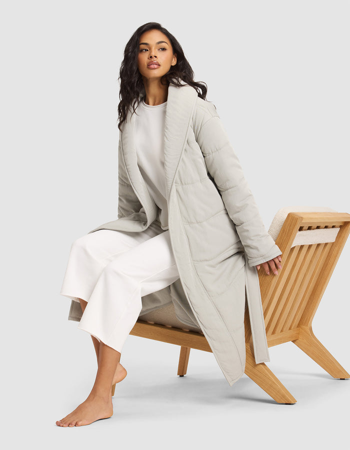 A woman with long dark hair sits barefoot on a wooden chair, wearing the Cozy Earth Women's Quilted House Coat over a white top and wide-legged white pants, against a plain light background. |Color:Aloe
