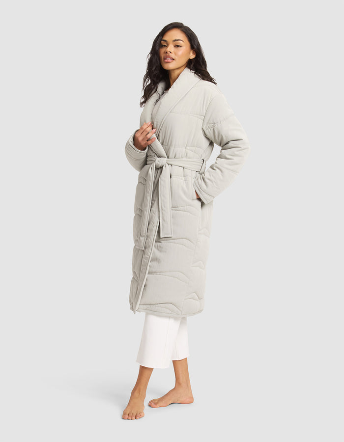 A woman stands barefoot against a plain light background wearing Cozy Earth's Women's Quilted House Coat, a long, light gray robe with a belt tie, styled over white pants. |Color:Aloe