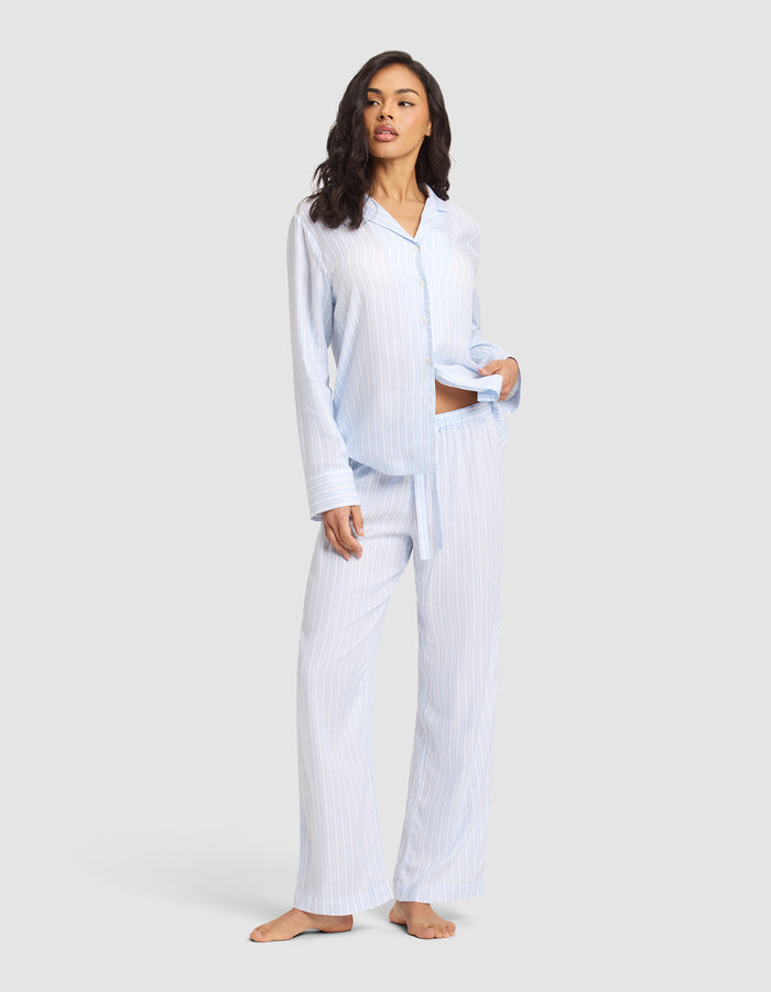 A woman stands barefoot against a light gray background wearing Cozy Earth Women's Soft Woven Pajama Pant in light blue and white stripes with a matching button-up shirt, her left hand in her pocket.  |Color:Spring Blue Stripe