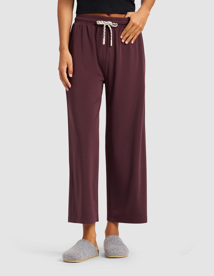 A person wearing Cozy Earth’s Women's Studio Cropped Wide Leg Pant in maroon, paired with a black top and gray slippers, stands against a plain light background.|Color:Raisin