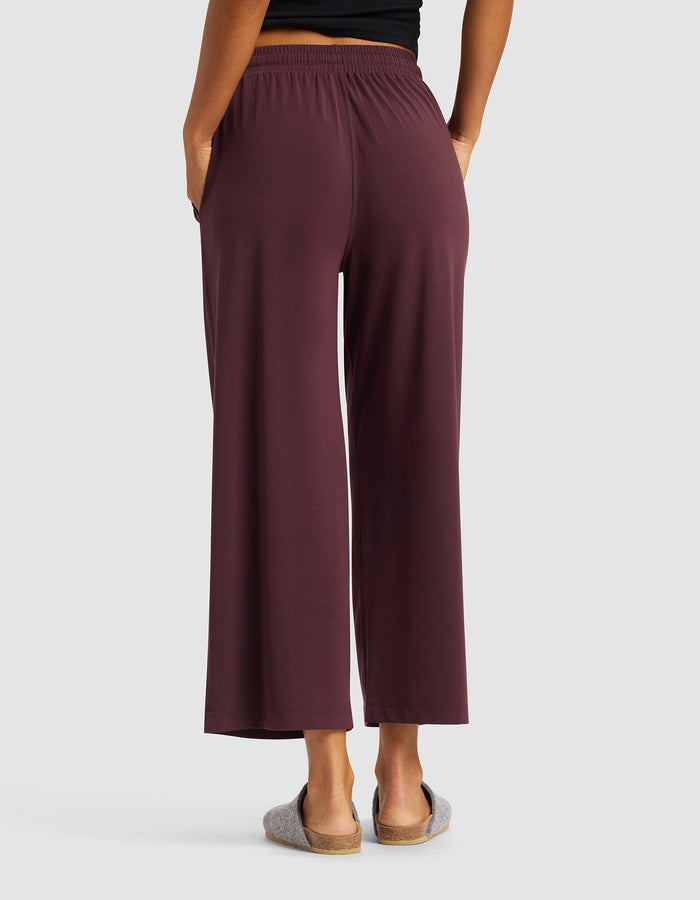 A person stands with their back to the camera, hands in pockets, wearing Cozy Earth's Women's Studio Cropped Wide Leg Pant in maroon, a black top, and gray slippers against a plain light gray background.|Color:Raisin