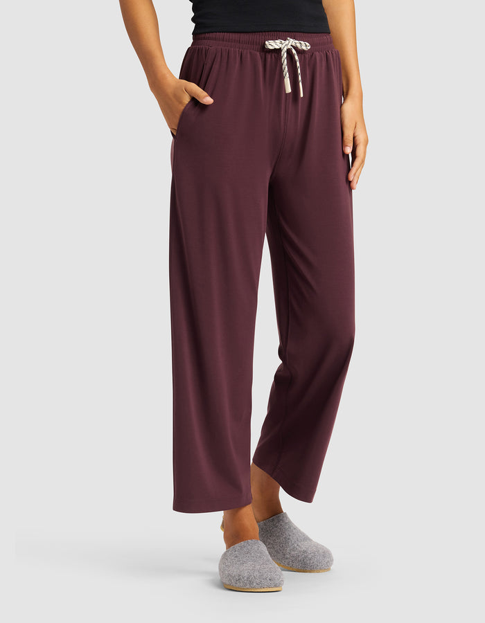 A woman models the Cozy Earth Women's Studio Cropped Wide Leg Pant in maroon with a white drawstring, paired with a black top and gray slippers, standing hands in pockets against a plain light background.|Color:Raisin