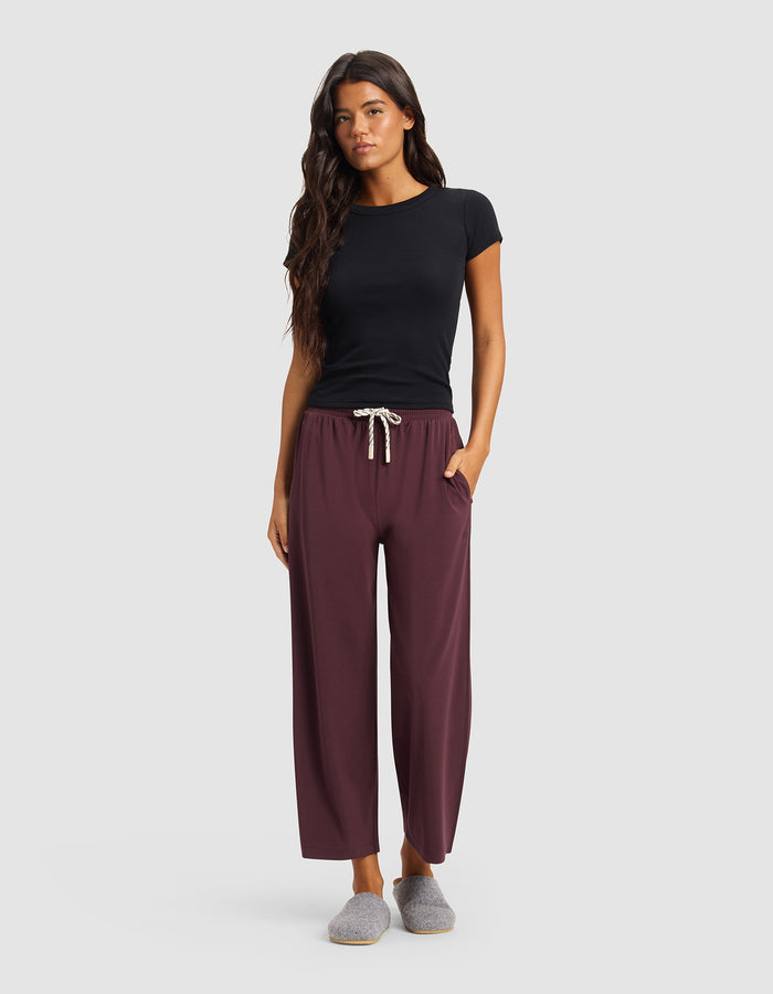 A woman faces forward against a plain light background, wearing Cozy Earth Women's Studio Cropped Wide Leg Pants in burgundy, a fitted black t-shirt, and gray slippers, with one hand in her pocket.|Color:Raisin