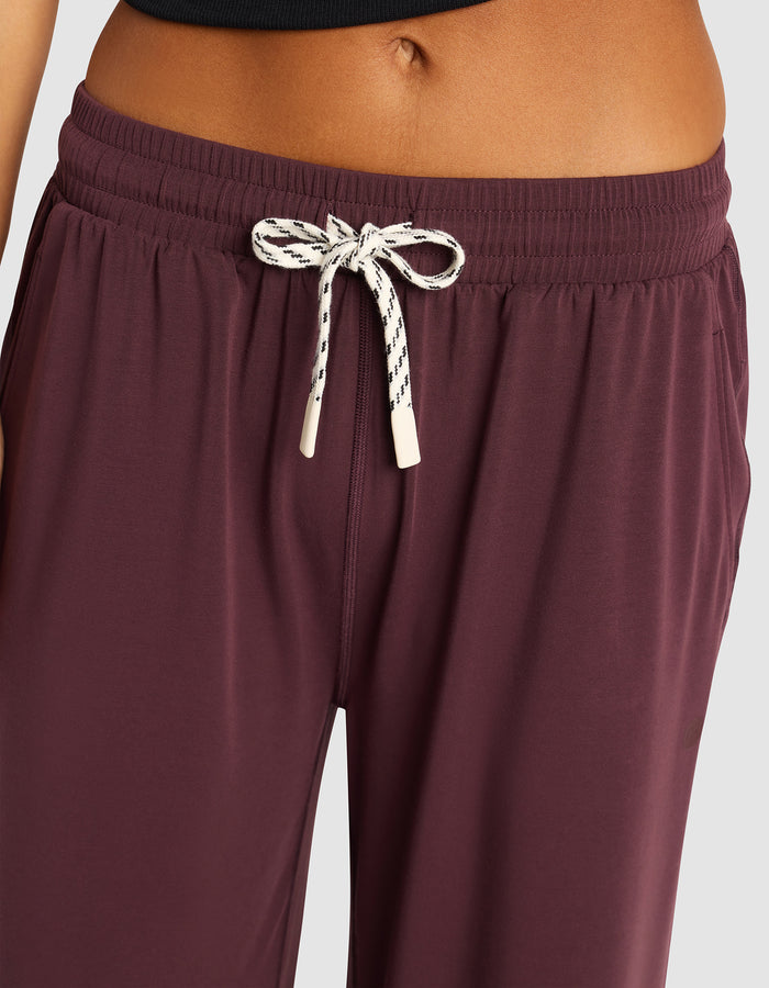 Close-up of a person wearing Cozy Earth Women's Studio Cropped Wide Leg Pant in maroon with a white and black tie at the waist, paired with a black top; the midriff and both arms are partially visible.|Color:Raisin