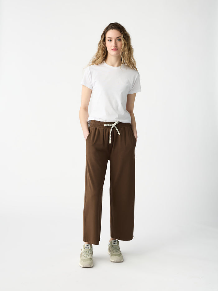 A person with long wavy hair stands against a white background, wearing a white T-shirt, Cozy Earth's Women's Studio Cropped Wide Leg Pant in brown with a drawstring, and beige sneakers. Their hands are in their pockets. |Color:Espresso