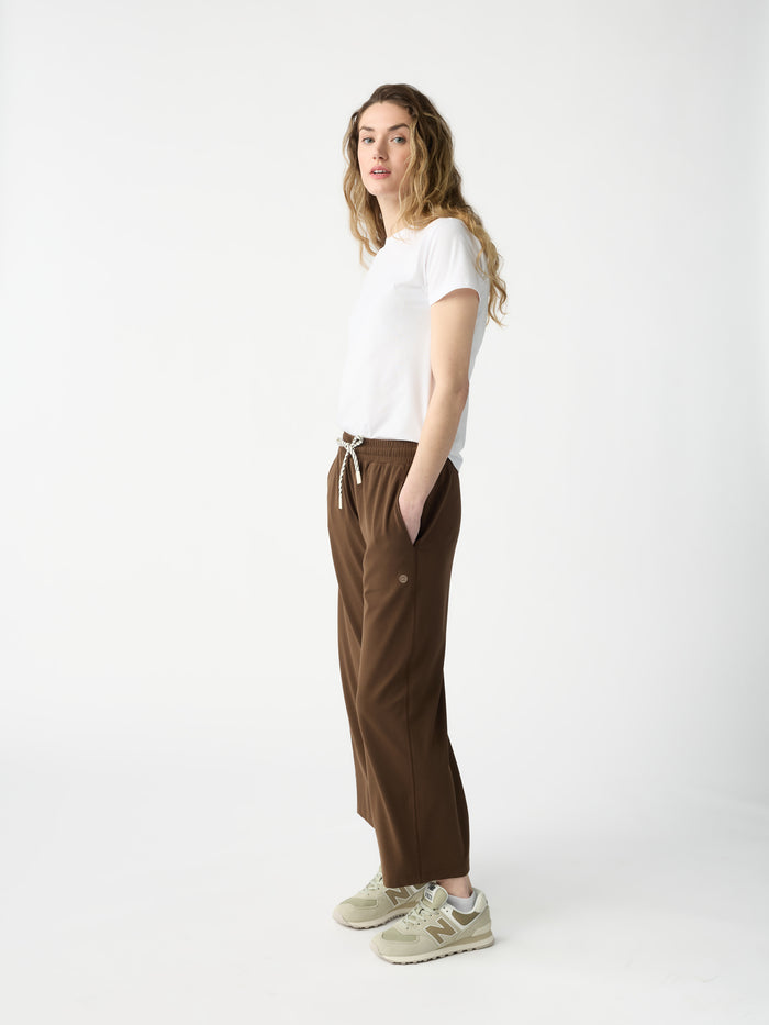 A person with long, wavy hair stands in profile against a white backdrop, wearing a white T-shirt, Cozy Earth's Women's Studio Cropped Wide Leg Pant in brown with a drawstring, and beige sneakers. Their hands are in pockets, and they have a neutral expression. |Color:Espresso