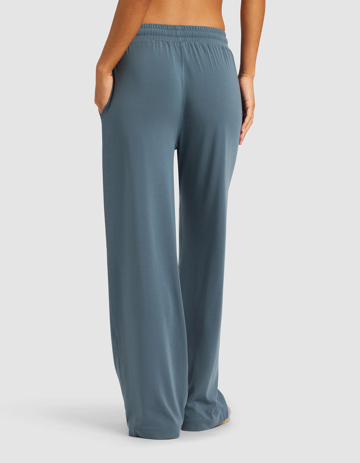 A person is shown from the back, barefoot against a plain background, wearing Cozy Earth’s Women's Studio Wide Leg Pant in loose-fitting blue-gray with an elastic waistband and side pockets.|Color:Dawn