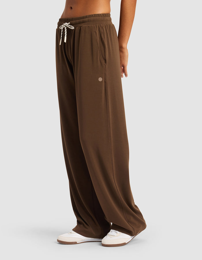 A woman wears Cozy Earth Women's Studio Wide Leg Pant—loose-fitting brown pants with an elastic waistband and white drawstring—paired with white sneakers, standing against a plain light gray background.|Color:Espresso
